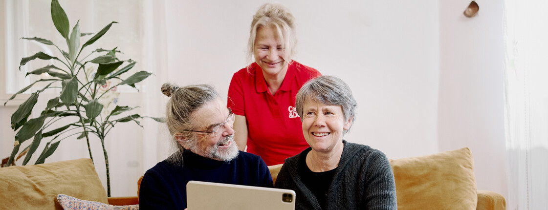 Drei Personen sitzen auf einem Sofa: Mann hält Tablet in der Hand, Frau daneben lächelt, Frau mit rotem Caritas Pflege Shirt steht hinter ihnen